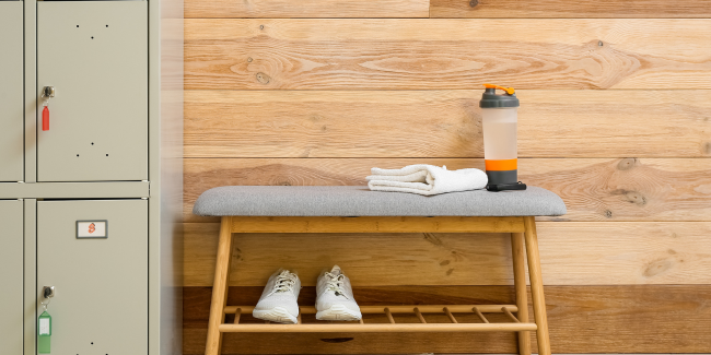 Visual image of a changeroom with lockers, bench, wooden wall backgroud with towel, water bottle on tne bench and shoes underneath