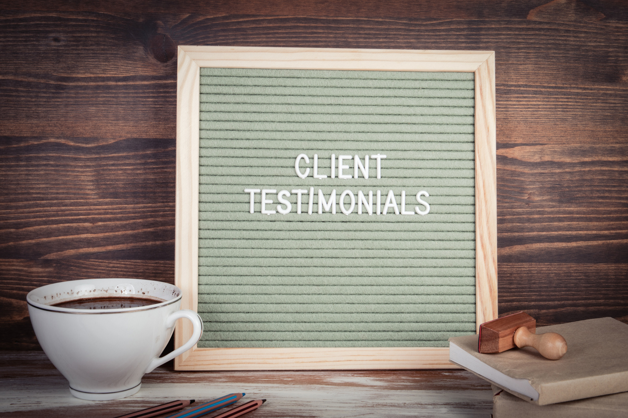 Letter board that reads Client Testimonials, sitting on a desk with a cup of coffee and notepad and pen